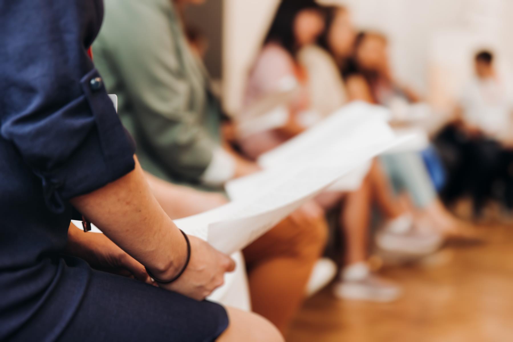 Nærbilete av hender som held eit ark. Fleire personar i bakgrunn. Bilete.