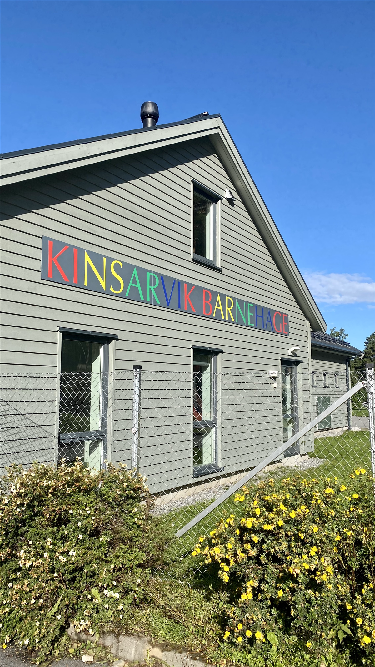 Bilete av hovudbygningen til barnehagen i Kinsarvik. Grått hus med fargerike bokstaver der det står Kinsarvik barnehage. Bilete. - Klikk for stort bilete