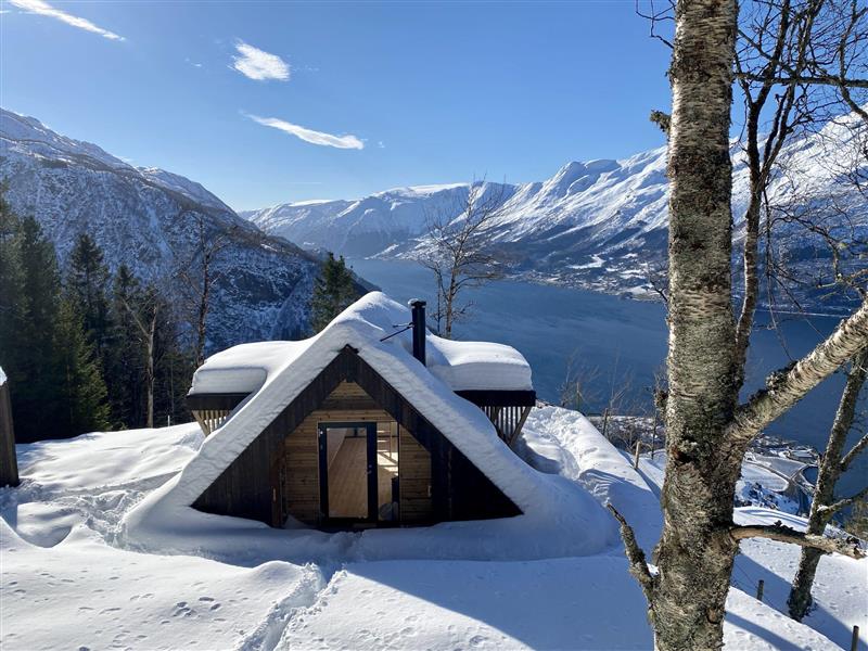 Dagsturhytta dekt med snø ein vinterdag. Utsyn over fjord og fin blå himmel. Bilete.
