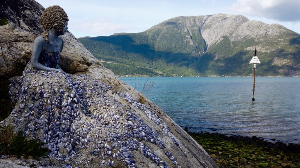 Bilete av havfrue i stein på Utne som skuer utover fjorden. Foto.